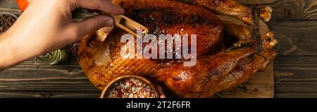 cropped view of man with wooden bowl seasoning grilled turkey for thanksgiving dinner, banner Stock Photo