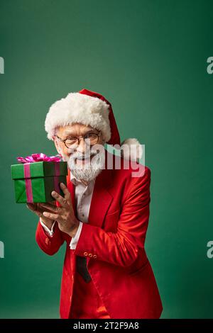 handsome stylish Santa in red hat and elegant attire showing present at camera, winter concept Stock Photo