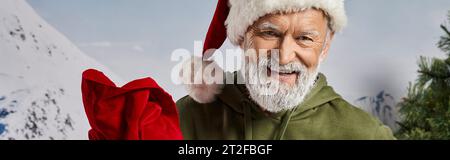 handsome Santa in Christmassy hat with red present bag smiling at camera, winter concept, banner Stock Photo