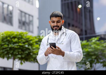 Arab smiling young male doctor, student standing near university hospital in uniform and using phone. Stock Photo