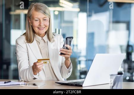 Senior smiling gray-haired business woman in a suit is working in the office on a laptop. Holds a phone and uses a credit card through an online application. Stock Photo