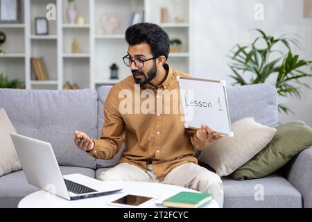 A young Indian male teacher teaches pupils and students remotely. He sits on the couch at home, shows and explains on the laptop camera the synopsis, the blackboard. Stock Photo