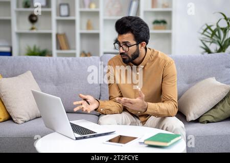 Indian young man businessman, freelancer holding a meeting online. Sitting on the couch and communicating via video call on a laptop. Stock Photo
