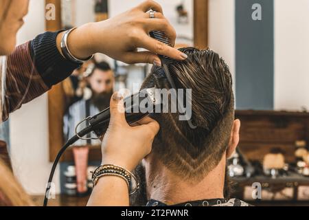 Hipster man at barbershop salon getting beard and hair cut - Hairdresser woman using hair clipper to modern cut with geometric drawing on the nape - B Stock Photo