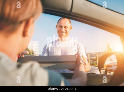 Young sincerely smiling deliveryman passing two pizza boxes to female sitting in car driver seat to on the city street. Small business, people relatio Stock Photo