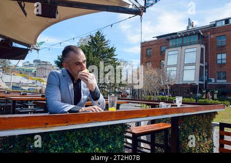 Caucasian, elegant adult man with jacket, short hair and business beard sitting outside restaurant eating, biting ham cheese sandwich and drinking ora Stock Photo