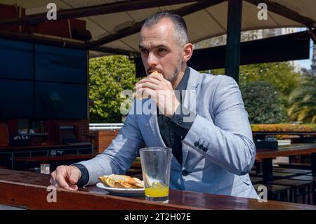 Caucasian, elegant adult man with jacket, short hair and business beard sitting outside restaurant eating, biting ham cheese sandwich and drinking ora Stock Photo