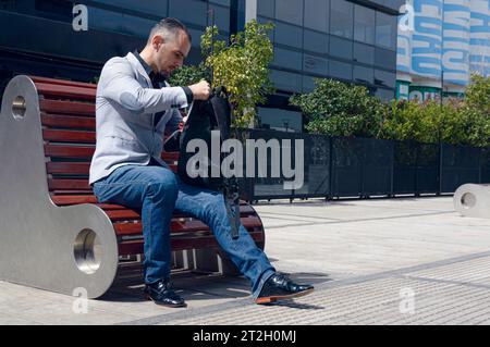 Caucasian adult businessman, sitting outside work buildings looking for something in his backpack, wearing stylish jacket and jean pants, business peo Stock Photo