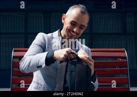 adult businessman, with short hair, beard and wearing stylish gray jacket, is sitting outside, laughing happily because he got money in backpack Stock Photo