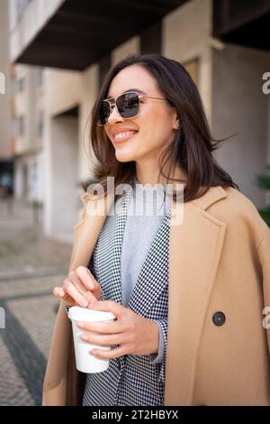 Streetstyle, street fashion concept: woman wearing trendy outfit walking in city. Cream trench coat, sunglasses. Side view Looking away, vertical photo Stock Photo
