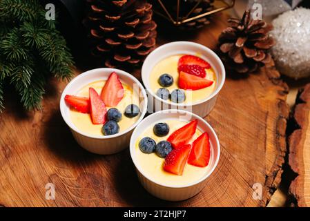 Three small servings of creme brulee with fresh blueberries and strawberries, pine cones, and a branch placed on a wooden background Stock Photo