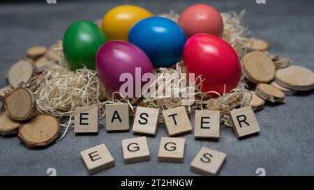Colorful eggs lie in a straw nest with wooden decoration around. On the table are letters that spell out the word "Easter Eggs". View from above Stock Photo