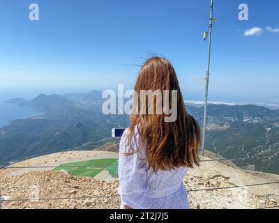 Hiker girl in casual clothes and with rbkzak takes pictures of natural mountain scenery with coniferous forest on smartphone camera, view from back. H Stock Photo