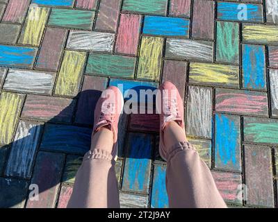 Beautiful female legs in pink sneakers and fashionable pants on a background of concrete paving slabs painted with joyful children's colorful bright c Stock Photo