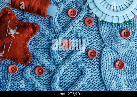 Beautiful texture of a soft warm natural sweater, fabrics with a knitted pattern and red small round buttons for sewing and a skein of thread, needle Stock Photo