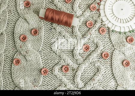 Beautiful texture of a soft warm natural sweater, knitted fabrics and red small round buttons for sewing and a skein of thread, needle bed. Flat lay. Stock Photo