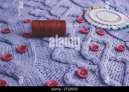 Beautiful texture of a soft warm natural sweater, knitted fabrics and red small round buttons for sewing and a skein of thread, needle bed. Flat lay. Stock Photo