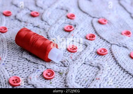 Beautiful texture of a soft warm natural sweater, fabrics with a knitted pattern of yarn and red small round buttons for sewing and a skein of red thr Stock Photo