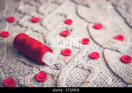 Beautiful texture of a soft warm natural sweater, fabrics with a knitted pattern of yarn and red small round buttons for sewing and a skein of red thr Stock Photo