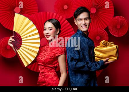 Smiling Asian couple in traditional costumes with fan and gold package as a gift for Chinese new year in oriental red background decoration Stock Photo