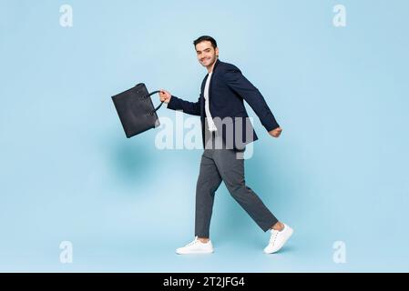 Full body happy male entrepreneur in smart casual clothes swinging arms with briefcase and looking at camera with smile while commuting to work agains Stock Photo