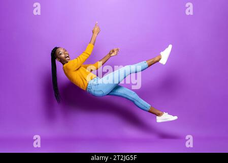 Full body side view of African American female in casual wear falling and levitating on purple background in studio Stock Photo