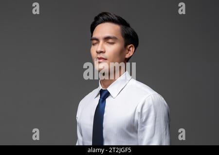 Handsome Asian businessman in white shirt with tie standing with closed eyes on gray isolated background in light studio Stock Photo