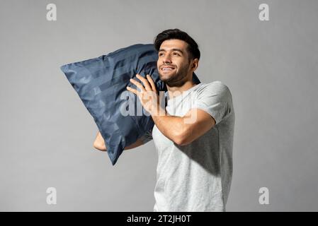 Sleepy Caucasian man with soft blue pillow standing on gray isolated  background in light studio Stock Photo