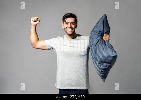 Positive Caucasian male with soft pillow showing muscles with smile on gray isolated background in light studio Stock Photo