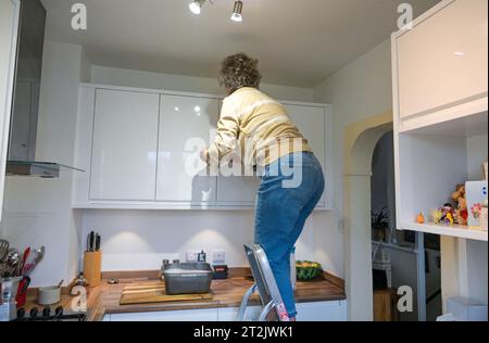Middle aged woman on a stepladder cleaning her kitchen cupboards with bowl of soapy water Stock Photo