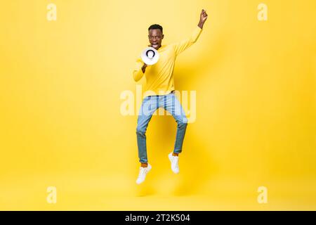 Powerful energetic young African man jumping and yelling on megaphone in studio yellow color isolated background Stock Photo