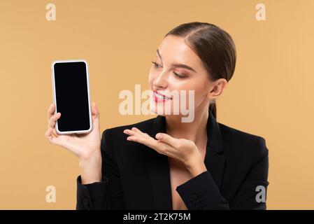 Beautiful Caucasian woman smiling and showing mobile phone with empty screen mock up in studio beige color isolated background Stock Photo