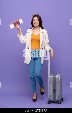 Beautiful young Asian woman tourist with baggage showing boarding pass and passport ready to fly isolated on purple background Stock Photo