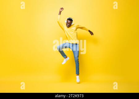 Ecstatic young African man wearing headphones listening to music and dancing with hand up in studio yellow color isolated background Stock Photo