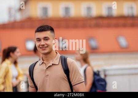 Close-up of a male portrait of a cute guy in a beige T-shirt with a black backpack. The teenager smiles and looks at the camera. Roofs of houses and o Stock Photo