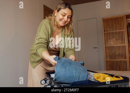 A young female blonde in a beige dress and a green jacket smiles and puts clothes in a suitcase in the bedroom in the house. An attractive woman in a Stock Photo