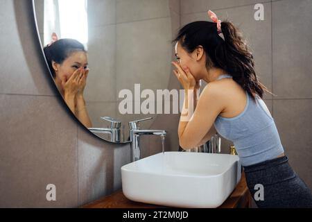 Young charming Asian woman in casual clothes washes her face over sink, looks at her reflection in round mirror. Cute Korean girl does water treatment Stock Photo