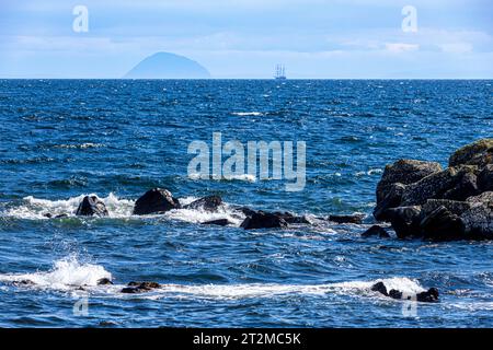 Waves breaking on rocks off the coast of the Kintyre Peninsula, with the barquentine 'Thalassa' (built 1980) approaching Aisla Craig , Argyll & Bute, Stock Photo