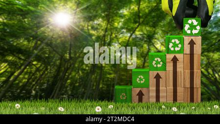 Gloved hand arranging wood blocks with green Recycling Symbols and brown arrows pointing upwards. On a green meadow with a blurry forest in the backgr Stock Photo