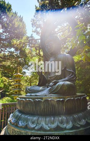 Sun beam of light hitting Buddha statue with palm raised and golden pagoda in background Stock Photo