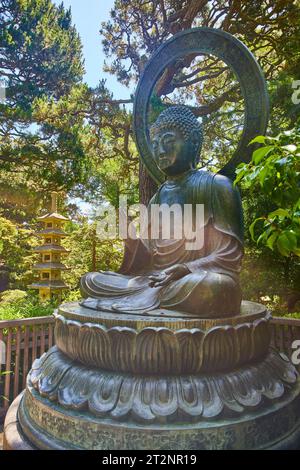 Sun flare lightly hitting Buddha statue with palm raised and golden pagoda in background Stock Photo