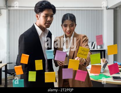 A professional man and woman stand together, engaged in a brainstorming session, using colorful sticky notes on a transparent glass wall. Stock Photo
