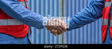 Two workers clad in reflective vests shake hands in front of a massive steel cargo container. Atmosphere at construction site. Concept of teamwork and Stock Photo