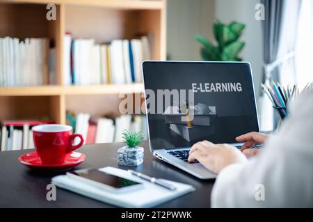 Young businesswoman sitting at table in front of laptop with inscription on screen e-learning and image of degree hat on books. Online education, e-le Stock Photo