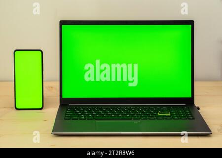 Photo of laptop and smartphone on wooden desk showing green screen. Stock Photo