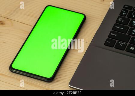 A smartphone with a green screen next to a gray laptop on a wooden table. Stock Photo
