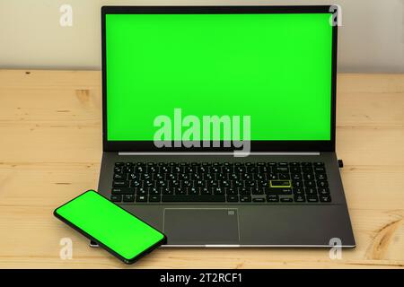 Photo of laptop and smartphone on wooden desk showing green screen. Stock Photo