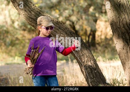 Teenage girl, dressed in comfortable suit, curiously collects firewood for fire while hiking in nature. Stock Photo