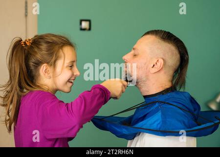 Young girl joyfully trims her father's beard at home, capturing a heartwarming father-daughter bonding moment. Stock Photo