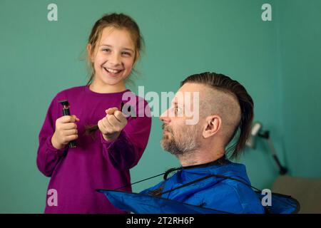 Daughter cutting her father's hair at home. The girl shows off the cut strands and laughs. Man is surprised Stock Photo